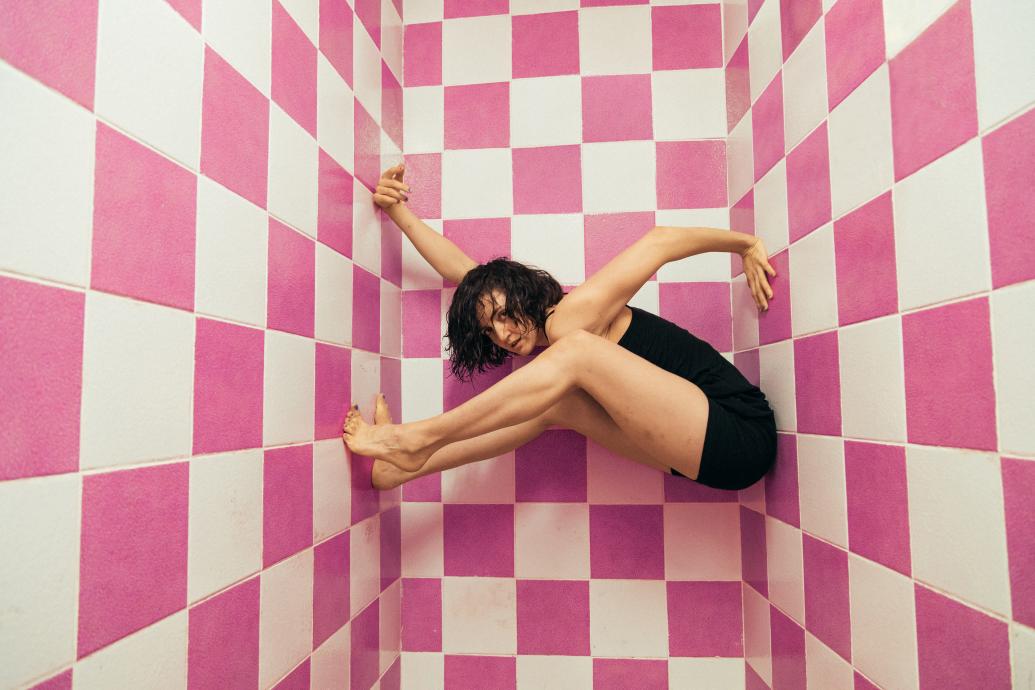 A woman in a black unitard holds herself suspended between pink and white tiled walls