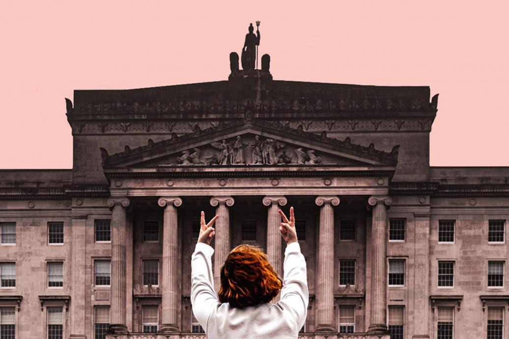 A woman with her back to camera faces Stormont, giving it two fingers up with both hands held up
