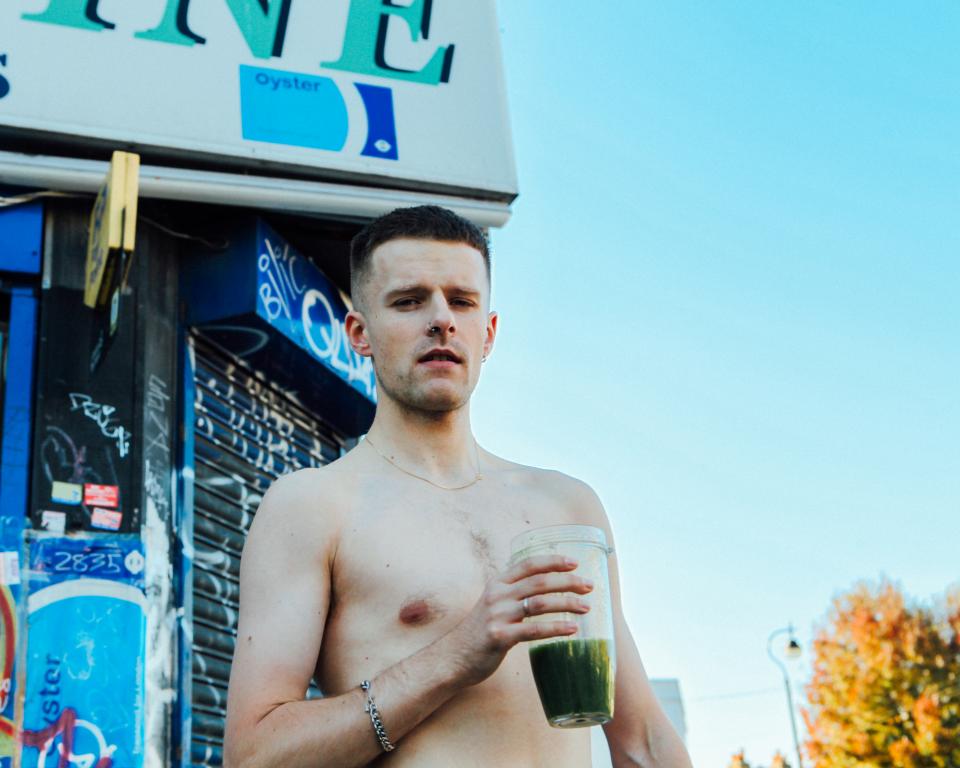 A topless man stands outside a shop during the day time holding a green smoothie