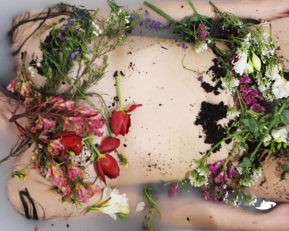 A woman's body floats in a full bath, her body covered in flowers and soil