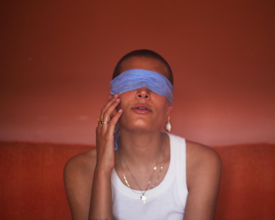A woman in a white top sits on a red sofa in a red room with a blue blindfold over her eyes and a hand to her face