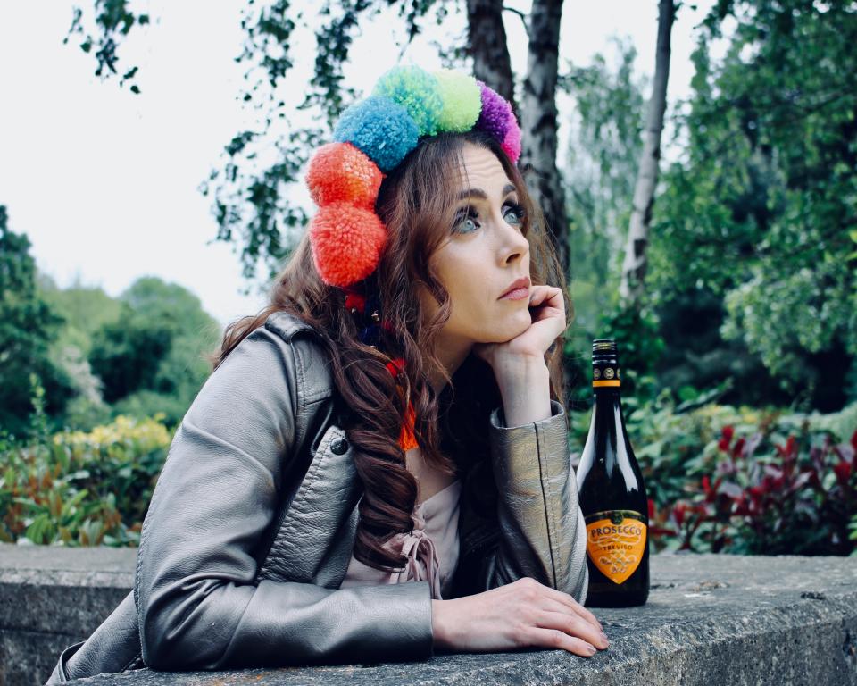 Modern Juliette in flower crown and biker jacket looking out wistfully from a balcony window, with a Buckfast beside her.