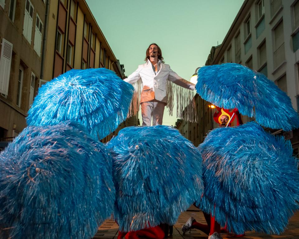 A man dressed in a white suit with fringed sleeves stands on top of a group of umbrellas covered in blue tinsel held by people wearing flame headdresses