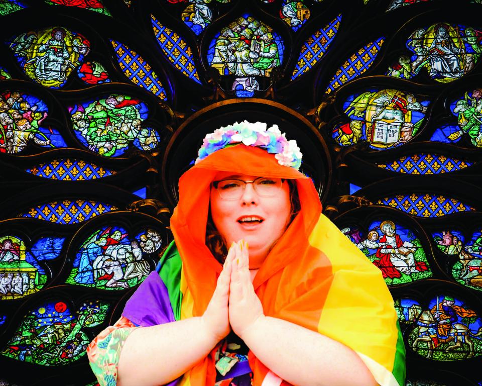 A woman poses against a stained glass window like the Virgin Mary, her hands held in prayer, her veil is a Pride flag and she wears a flower crown
