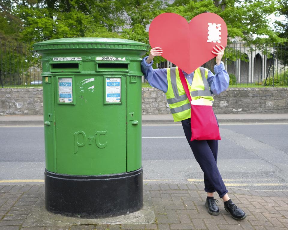 Postwoman holding a heart envelope beside an Irish postbox