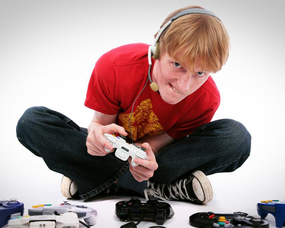 A man sits on the ground in a white room with a headset on, surrounded by game controllers on the floor, as he holds one in both hands