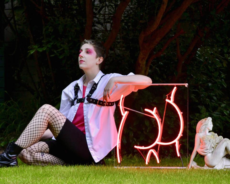 Púcaparty Glitter Hole, A woman sits on the grass leaning against a neon sign in a pose like the White Lady of Cabra statue sitting behind her