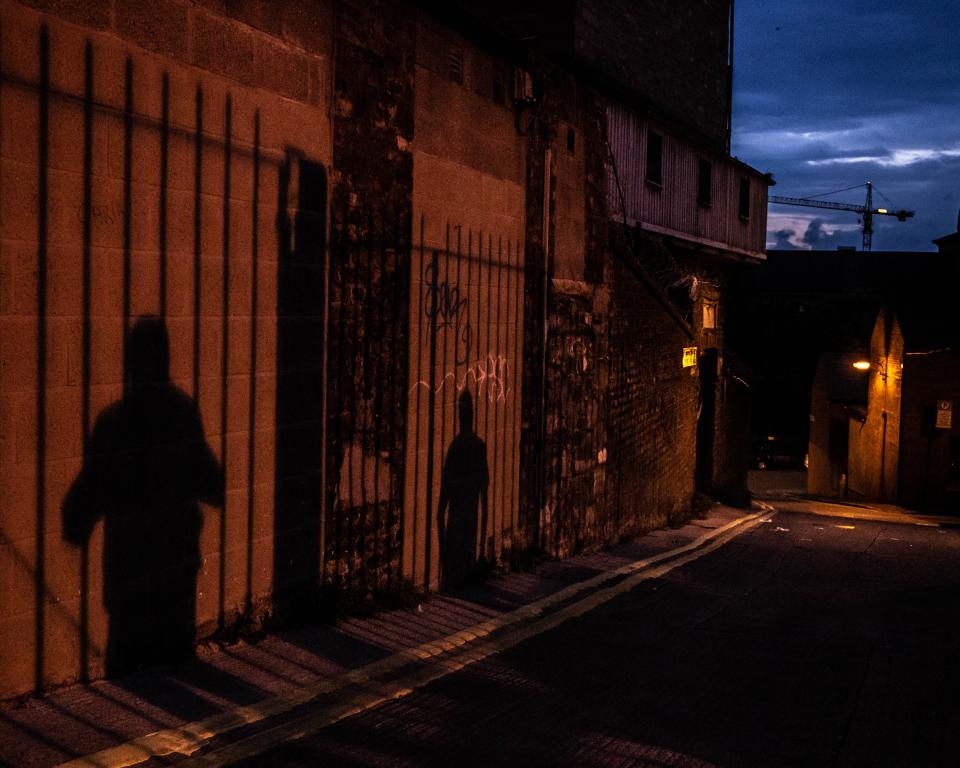 The shadows of two people, standing apart against railings, are projected onto an alley wall at night