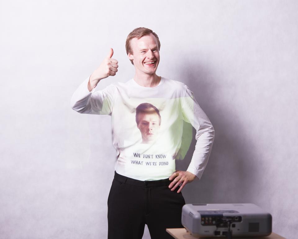 A man smiles, giving a thumbs up, as an image of himself frowning and holding a sign saying "We don't know what we're doing" is projected onto his top