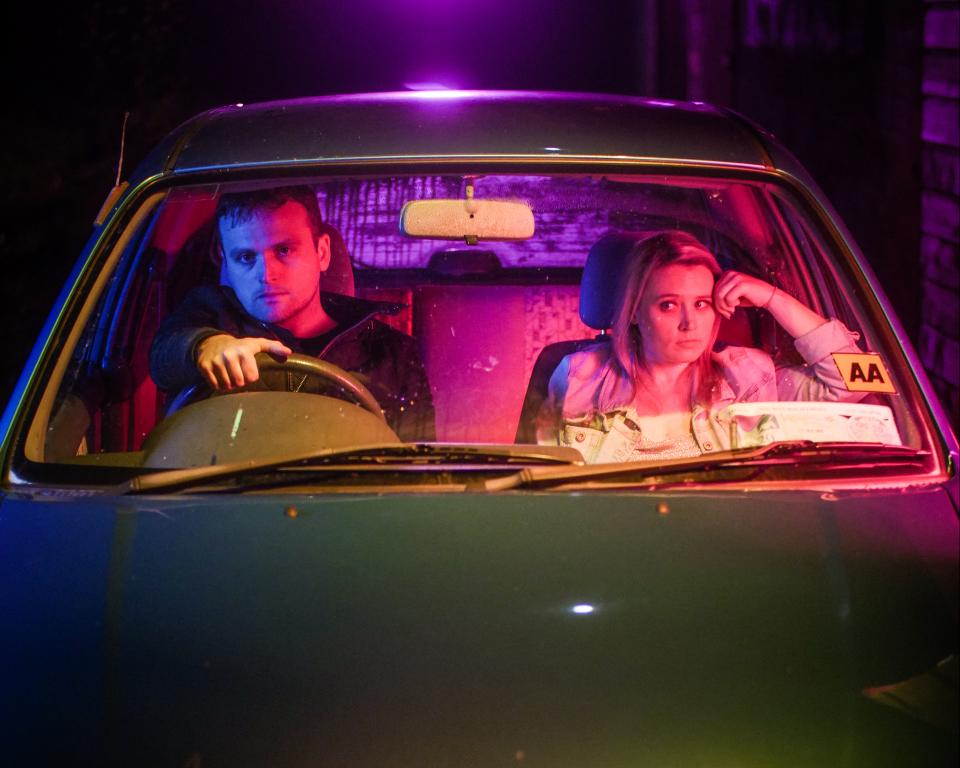 A man sits in the driving seat and a woman in the front passenger seat of a car awkwardly at night time