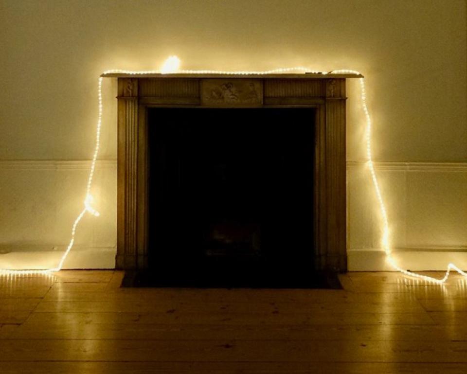 A dark fireplace is draped with a line of fairy lights that continues across the floor on either side of it in a beige room with wood floors
