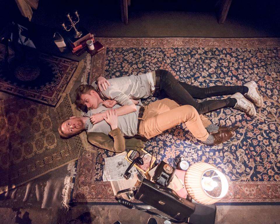 Two men lie together on a carpet holding each other