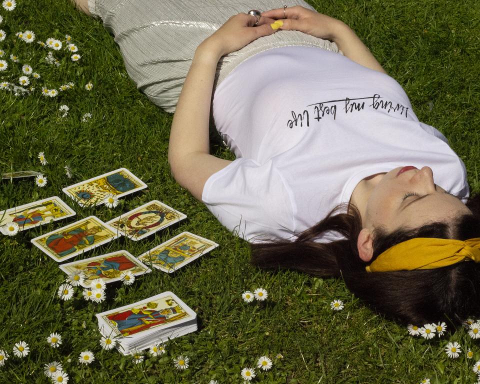 A woman lies on the grass surrounded by daisies with a deck of tarot and a tarot spread laid out next to her