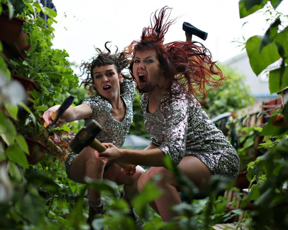 Two fierce women in sequin dresses crouch with garden weapons in leafy surroundings