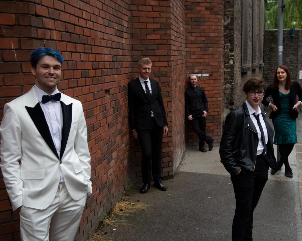 5 well dressed people stand along a red brick wall