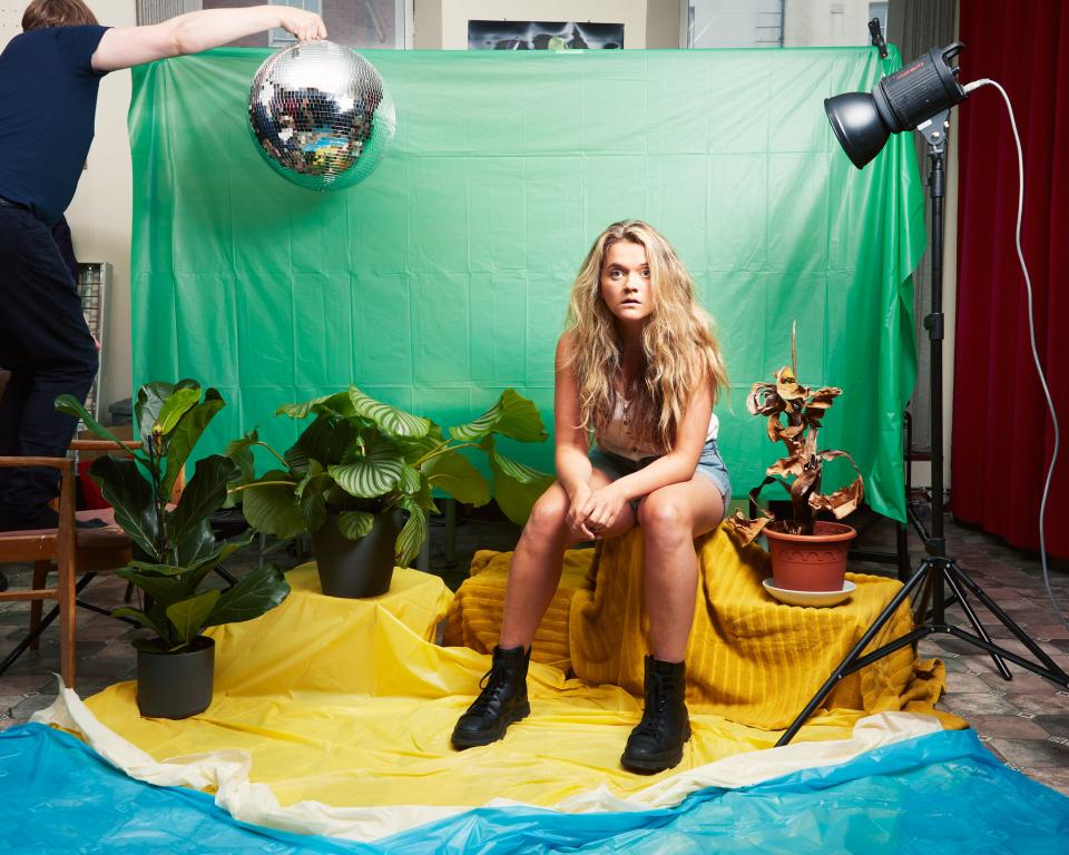 Woman sitting by green screen surrounded by props of plants and a disco ball held in the air by a man