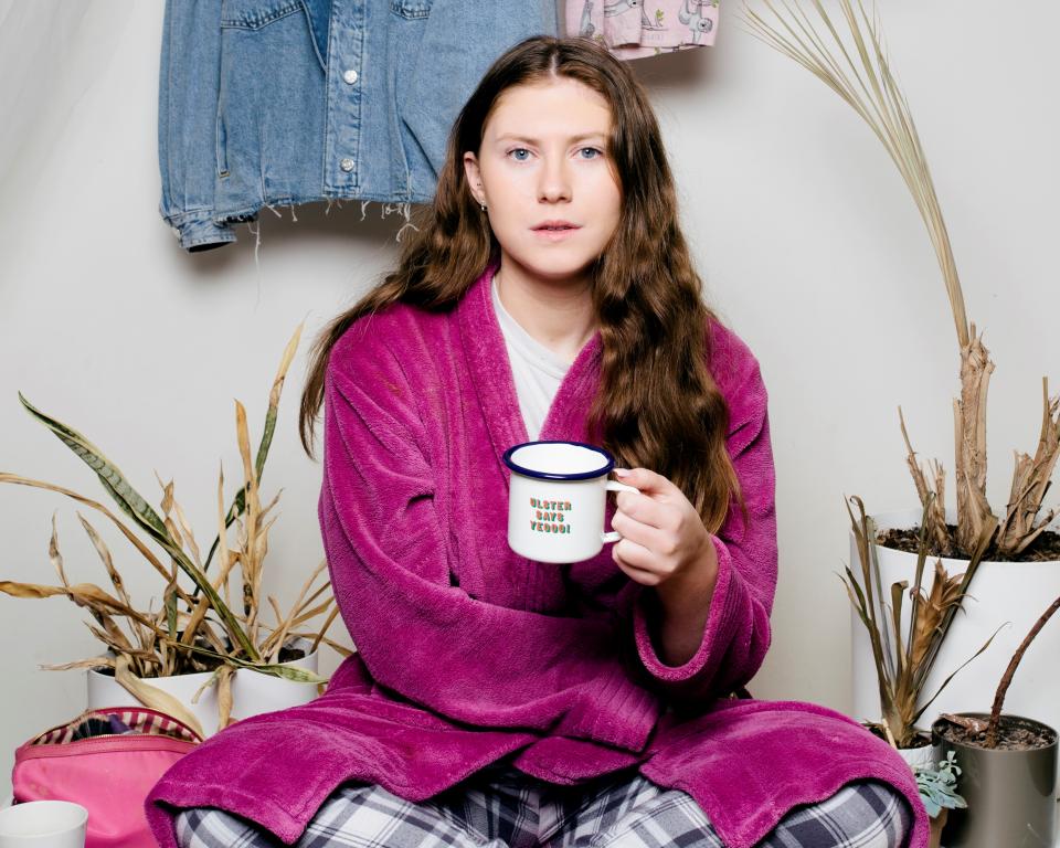 Young woman seated with a cup of tea surrounded by clothes and dead plants