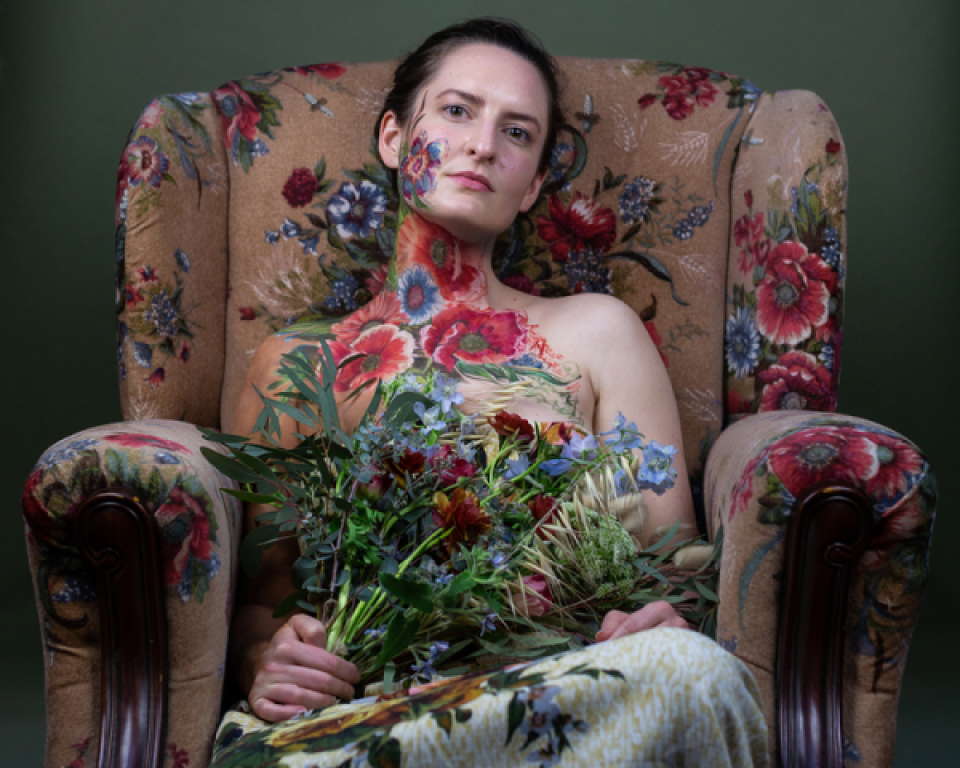 A woman with a floral design painted on her skin with flowers sitting in a floral upholstery armchair