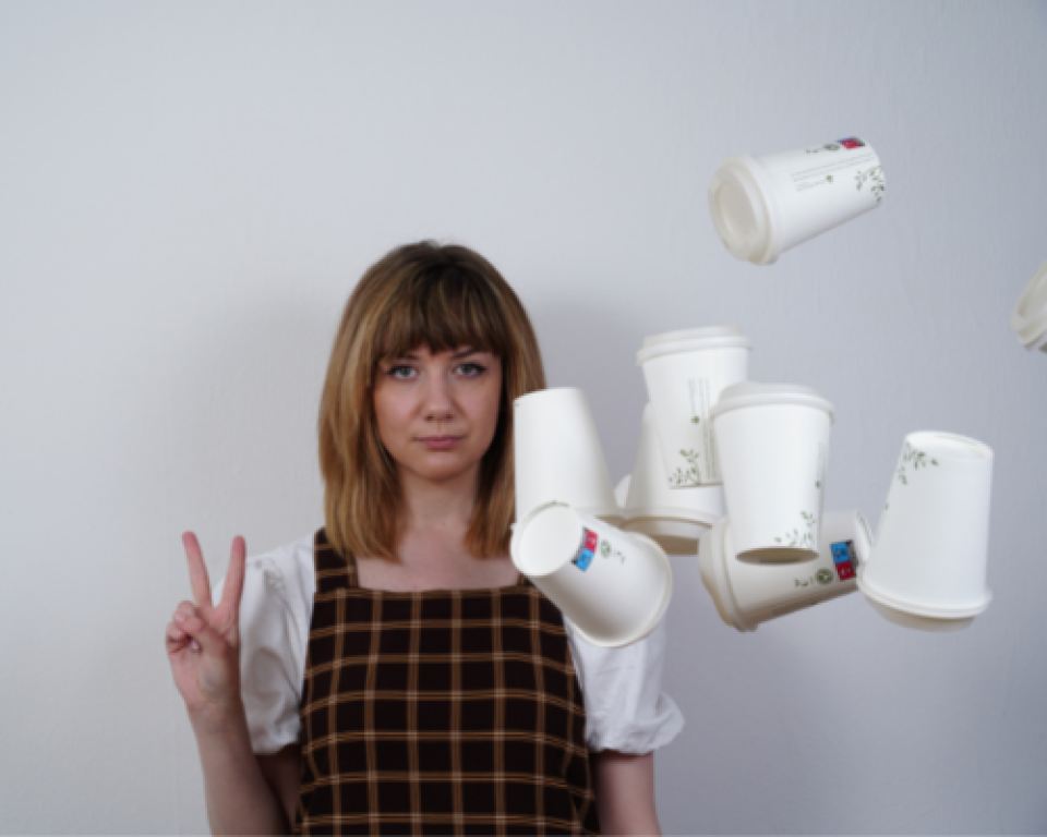 A woman stands against a wall holding up a peace sign while coffee cups come flying at her face