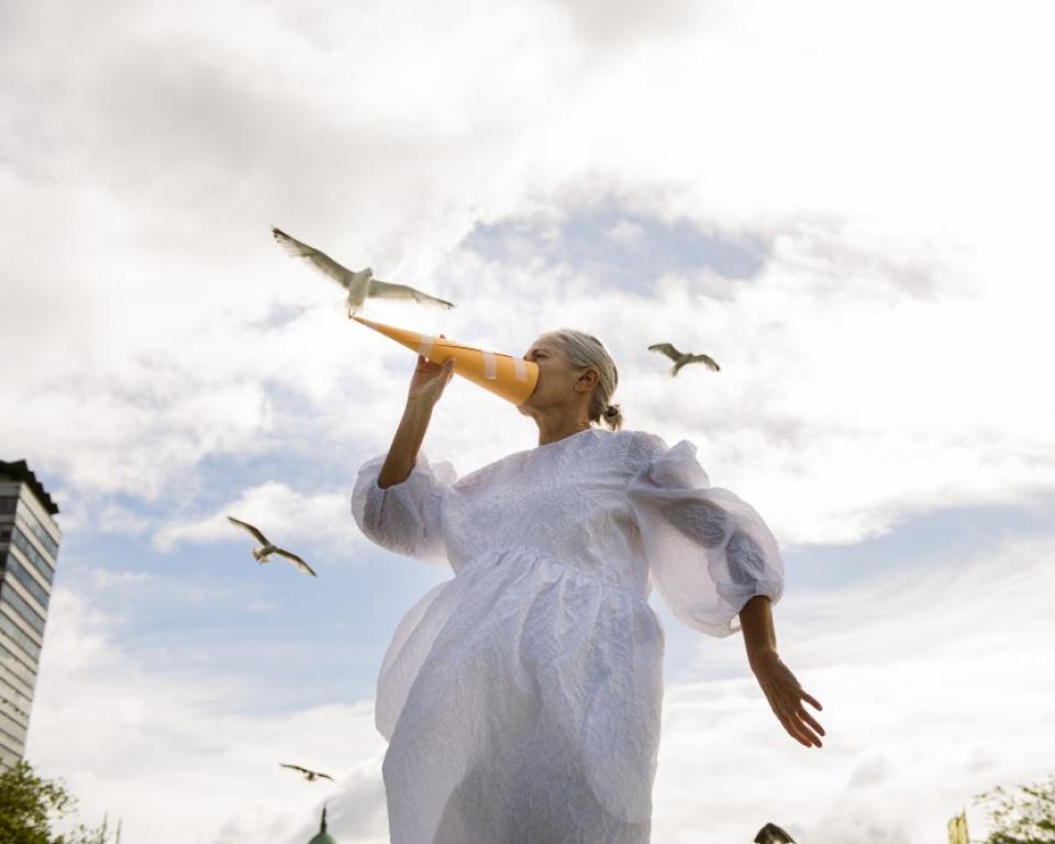 A woman in a white dress holds a paper beak to her face against a cloudy sky in the background