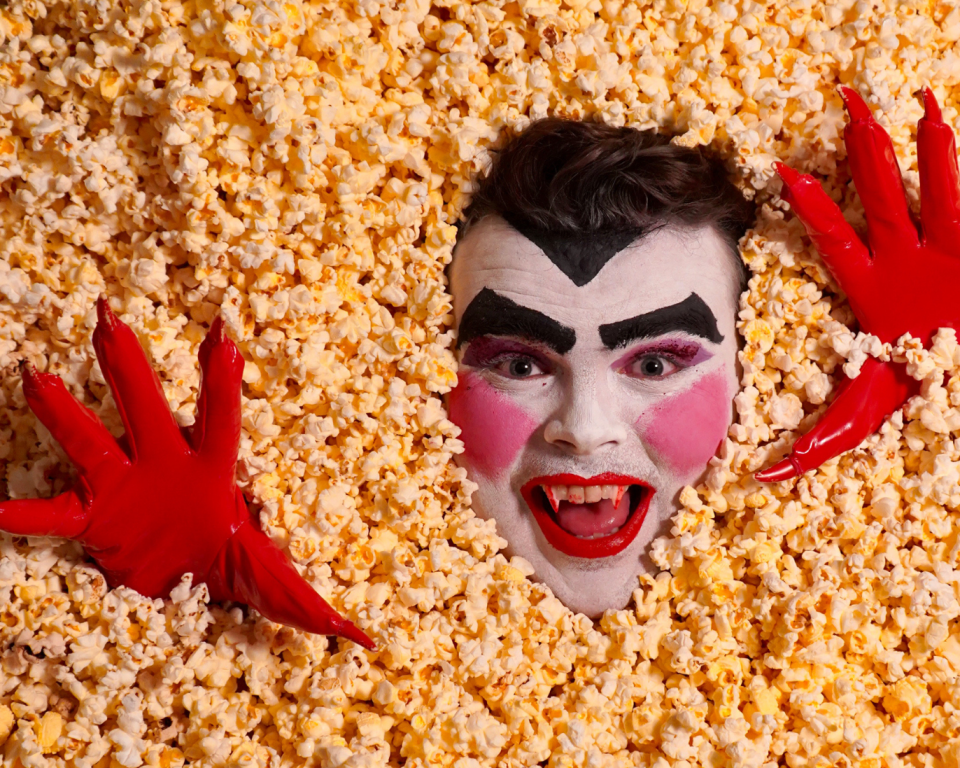 A face painted as a vampire and plastic red gloved hands appear above a layer of popcorn