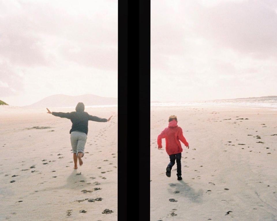Two people running on the beach, one capturing the moment with a camera while enjoying the sunny day.