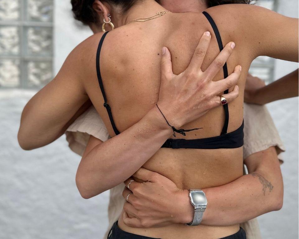 Two women embrace each other while wearing matching black bikinis, smiling joyfully in a sunny outdoor setting.