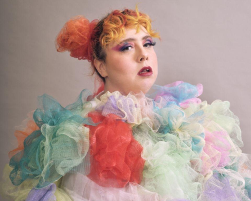 A woman with vibrant, multicolored hair wearing a bright, patterned dress stands confidently, showcasing her unique style.