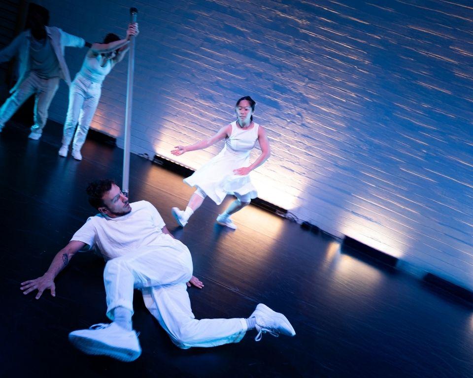 A man in white lies on the floor surrounded by other dancers in motion, showcasing a dynamic dance performance.