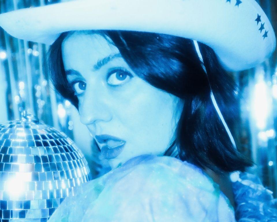 A woman wearing a cowboy hat stands beside a shimmering disco ball, blending country and disco themes.