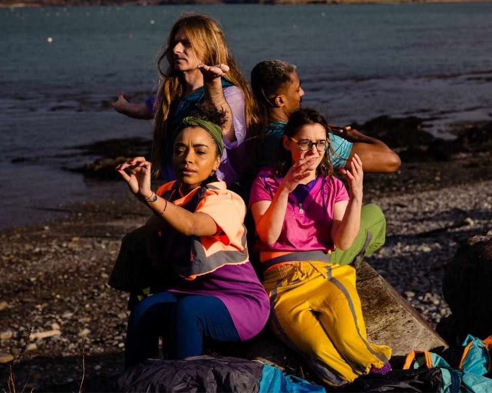 Three women in vibrant outfits sit on rocks by the water, enjoying a sunny day and the scenic view around them.