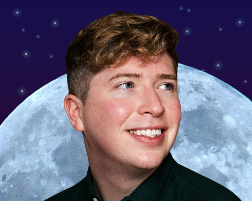 A man poses behind a moon, looking away from the camera and smiling 