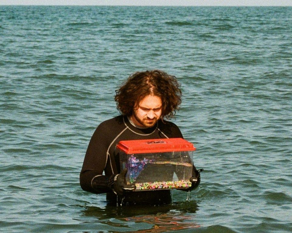A man in a wetsuit holds a fishtank while standing in the water, looking focused and engaged in an activity.