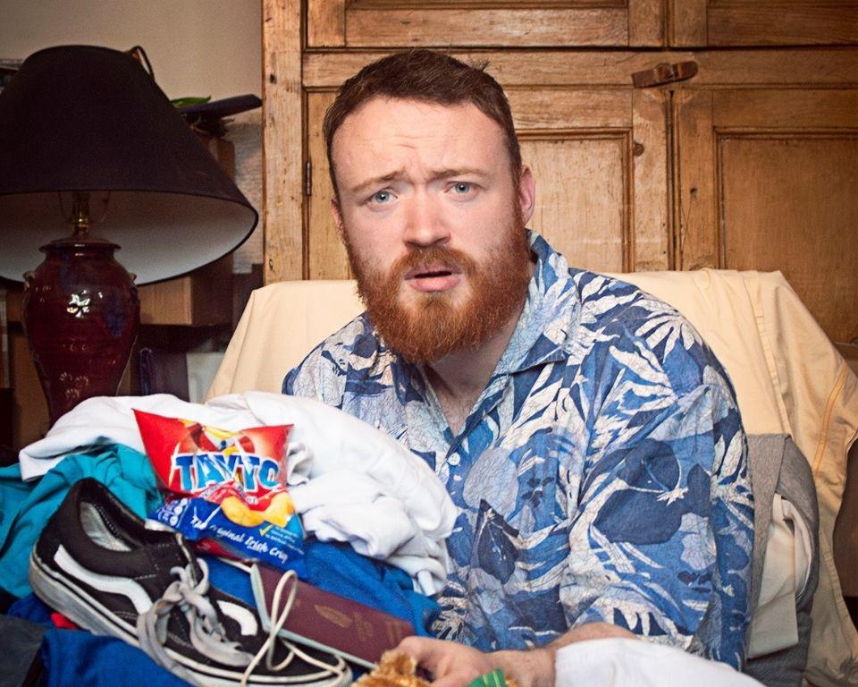 A man with a beard wearing a blue shirt sits in a chair next to a large pile of clothes.
