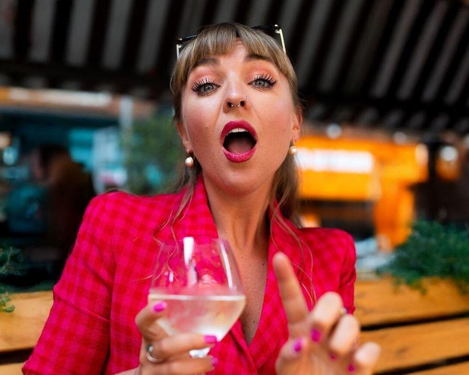 A woman in a red jacket raises a glass of white wine, outside a bar.