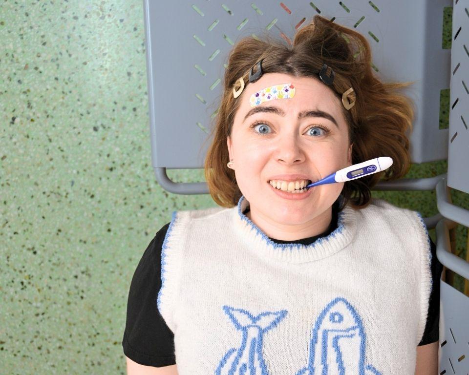 A person lies down in a medical setting, wearing a light sweater with fish designs, while a thermometer is positioned near their mouth.