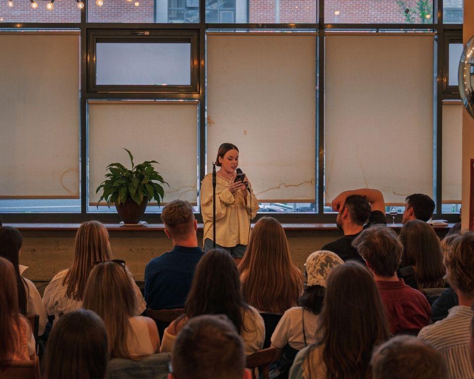 A speaker stands on stage with a microphone, engaging an attentive audience in a cozy, well-lit venue.