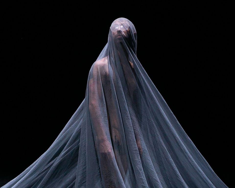A figure stands shrouded in a sheer gray fabric, partially obscuring their body and face, against a dark background.