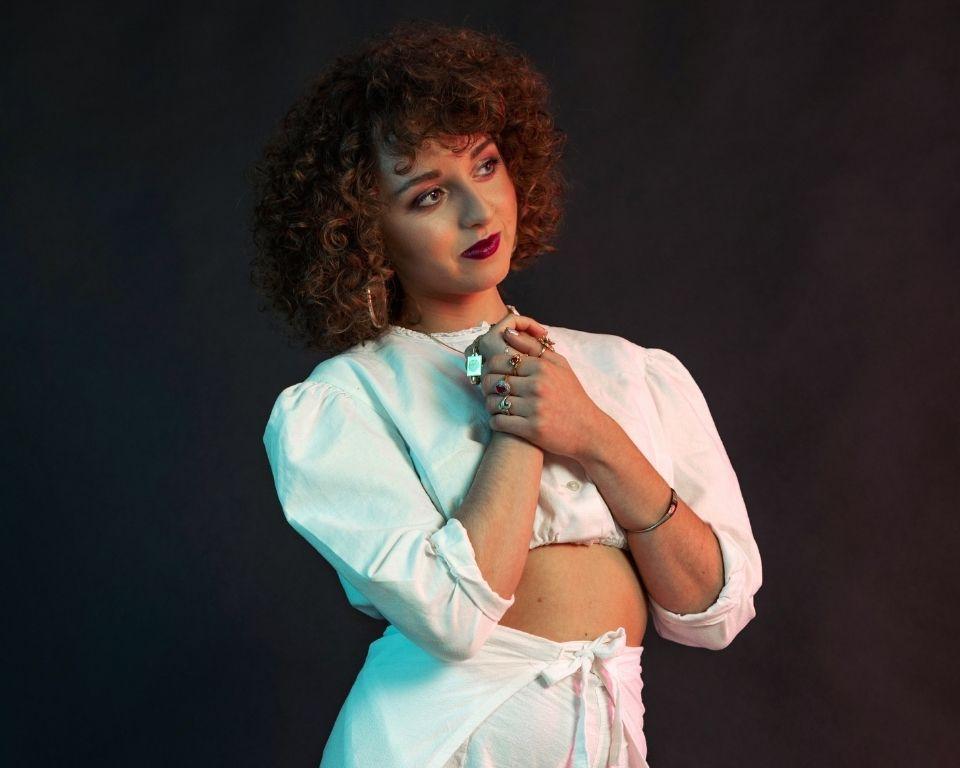 A person with curly hair poses gracefully in a stylish white outfit, showcasing intricate jewelry against a dark, moody background.