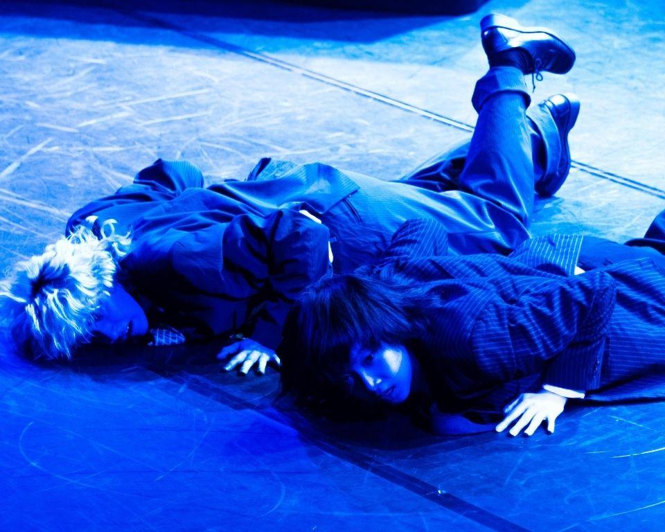 Two performers in formal attire lie on a dark stage, illuminated by blue lighting.