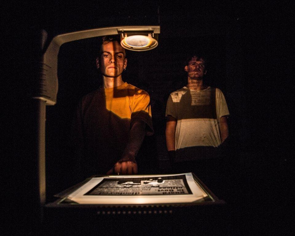 Two figures stand in dim lighting, one casting a shadow on a lightbox displaying an illustrated document.