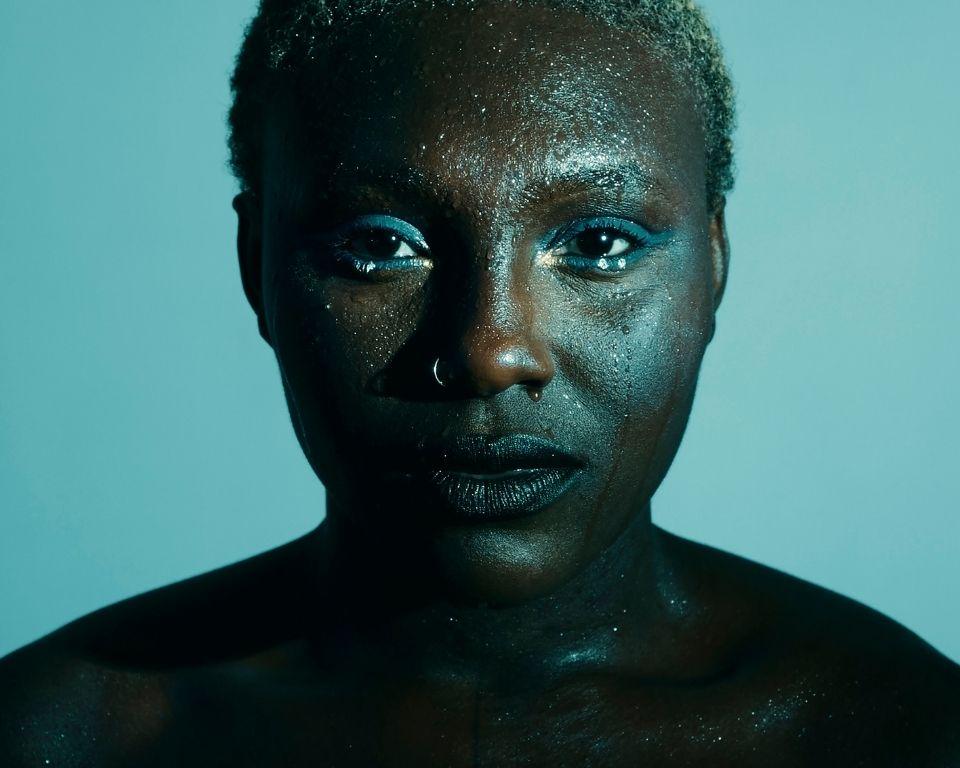 A person with black skin wearing blue eyeshadow, with prominent cheekbones, captured in a portrait shot indoors.