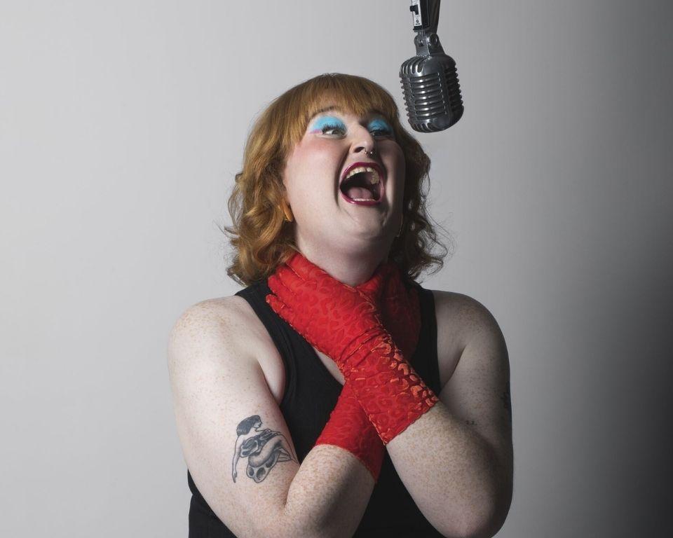 A woman singing into a hanging microphone, wearing lipstick and fashionable clothing, with her shoulder and elbow visible as she holds her neck
