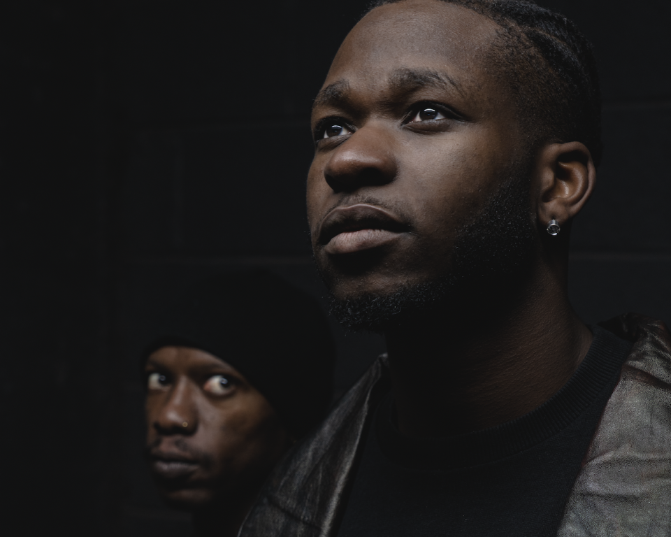 Two men standing next to each other against a black background, with one man staring at the other intensively