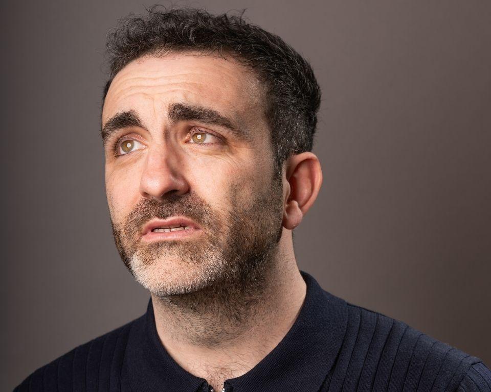 A bearded man dressed in a blue shirt, standing against a plain background with a calm demeanor.