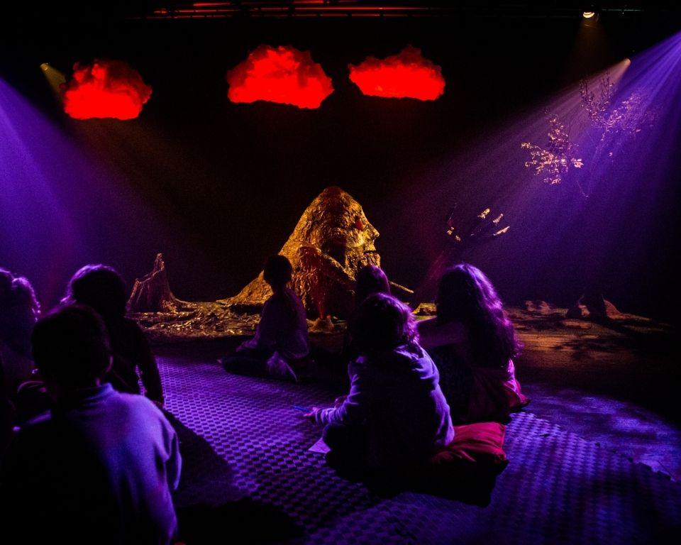 A group of children sit cross-legged on cushions in a dimly lit theatre space, watching an abstract, nature-inspired performance. The stage features a glowing central figure made of textured materials, resembling a mountain or creature, with dramatic purple and red lighting. Red clouds hang above, and a tree stands to the right, creating a dreamlike, immersive atmosphere.