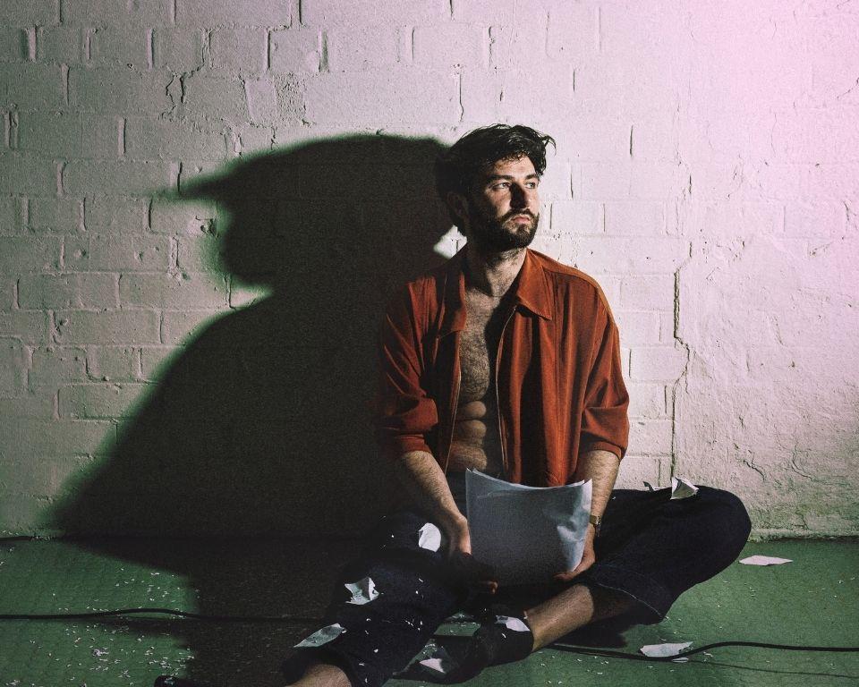 A man on the floor, holding a piece of paper but looking thoughtfully away from it.