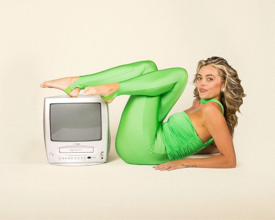 A woman with neon green top and legging, laying on the floor with her feet on a tv in an 80s pose.