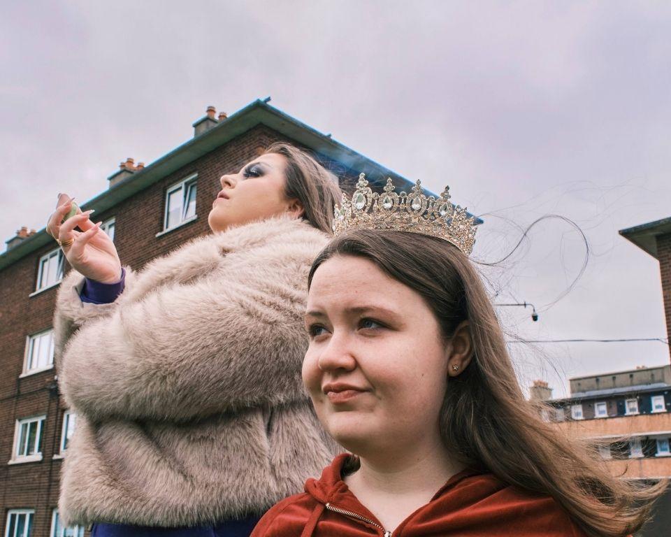 A young girl wearing a tiara stands in the foreground, while a woman in a fur coat vapes in the background, with residential buildings behind them under a cloudy sky.