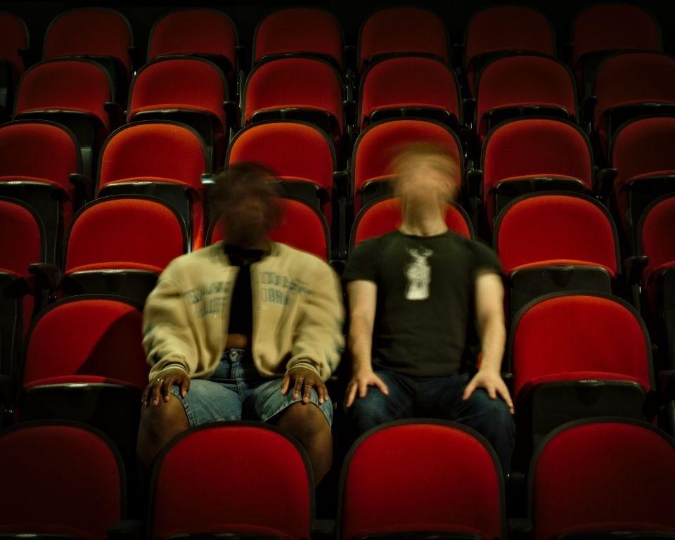 Two people sit side by side in the middle of an empty theatre with red and black seats. Their faces are blurred in motion, creating a ghostly, disorienting effect. One wears a light jacket and shorts, the other a black t-shirt. The lighting is low, adding to the eerie, surreal mood.